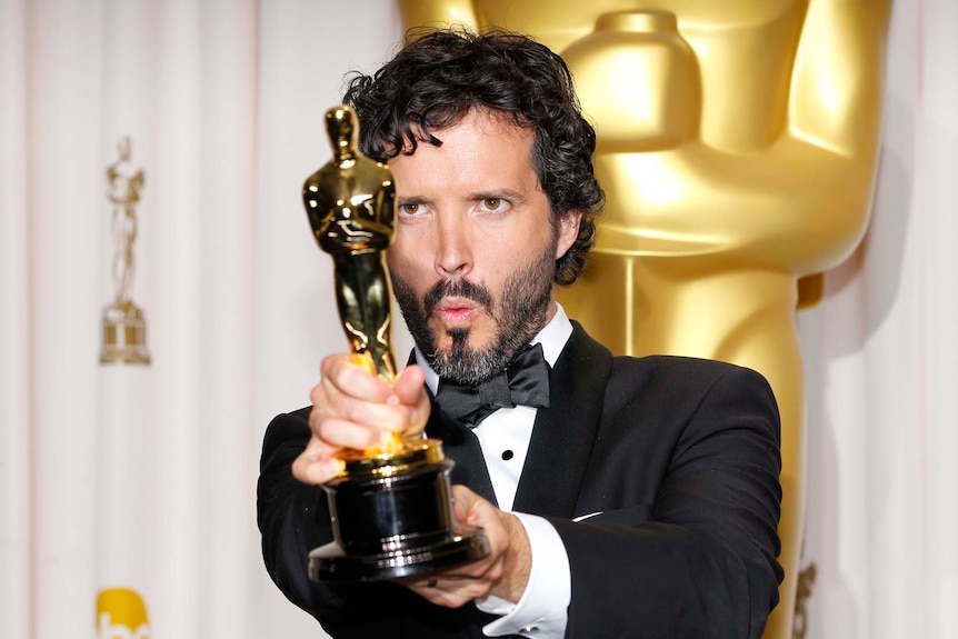 Bret McKenzie holds his Oscar, makes a face, wears black tie. A large oscar statue behind.