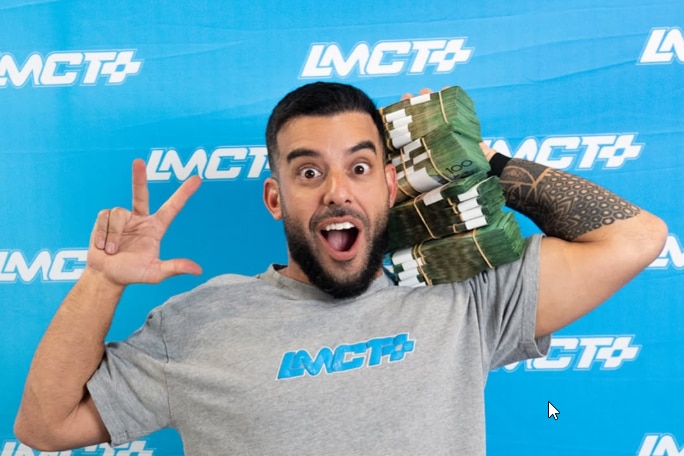 A man in front of a blue background holding a large amount of $100 bills on his shoulder.