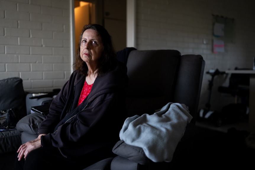 Lori sits in a dimly lit, small apartment living room with a withdrawn expression.