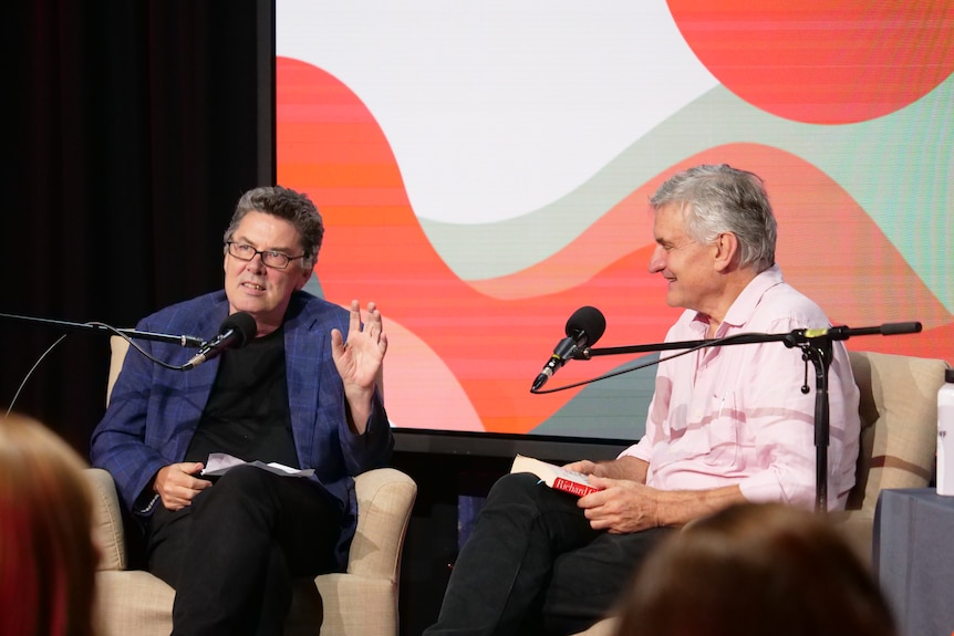 Two men sit on separate chairs with microphones in front of them.