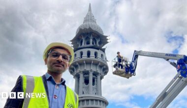 A man wearing a white safety helmet and tabard with the top of an ornate grey tower in the background.
