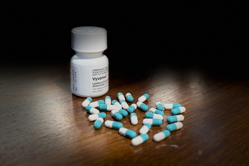 White and blue pills are scattered on a wooden table with a white screwcap bottle in the background.