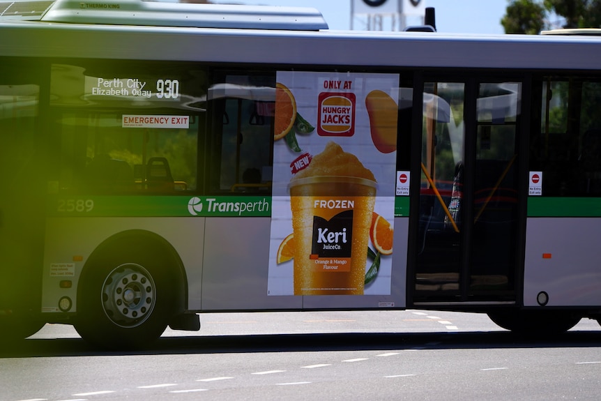 A junk food advertisement for an orange drink from Hungry Jacks on the side of a Transperth bus as it travels along a road.