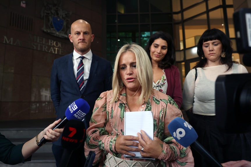 Microphones point at blonde woman in a peach and green dress holding a piece of paper as people stand behind her.