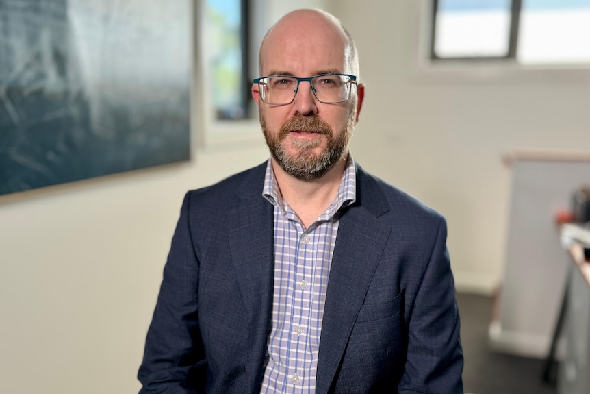 A bald caucasian man wearing glasses, a chequed shirt, and blue blazer jacket looks on with eyebrows slightly scrunched