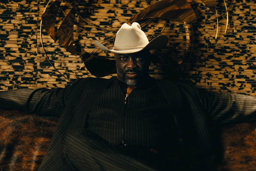 Adewale Akinnuoye-Agbaje in character as Alamo dressed in a cowboy hat sitting down with arms out wide