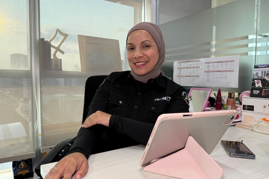 Woman wearing hijab sitting behind her desk smiling.