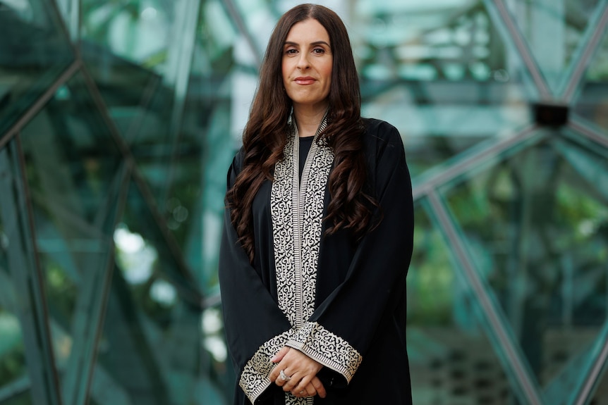 A woman with long brown hair, wearing a black embroidered jacket, with her hands clasped together.