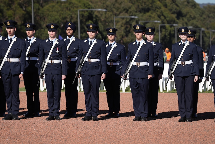 A row of military people in blue with firearms stands in a uniform row on gravel.