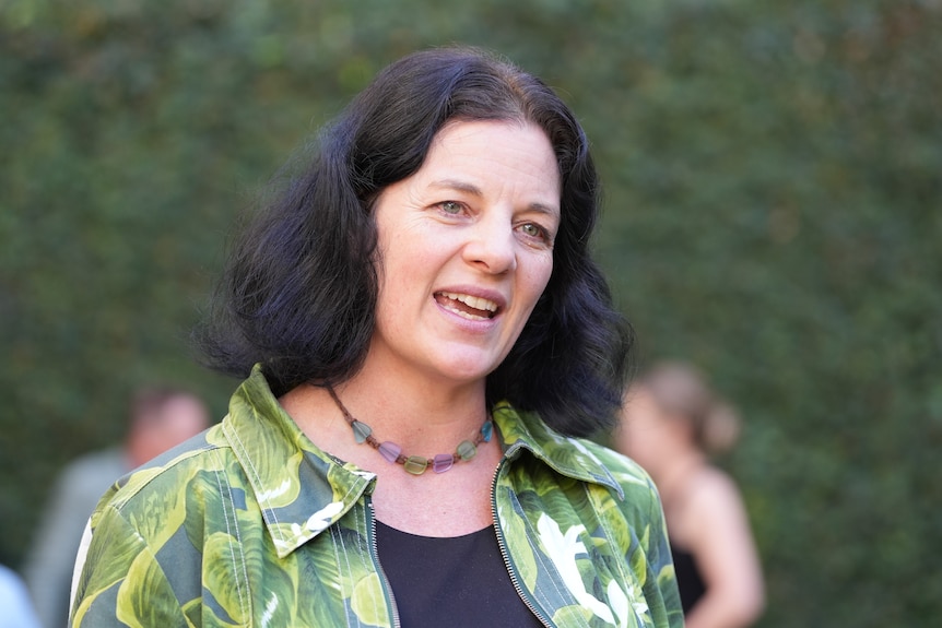 A woman with dark hair and a green blouse speaks, standing outside near a green hedge.