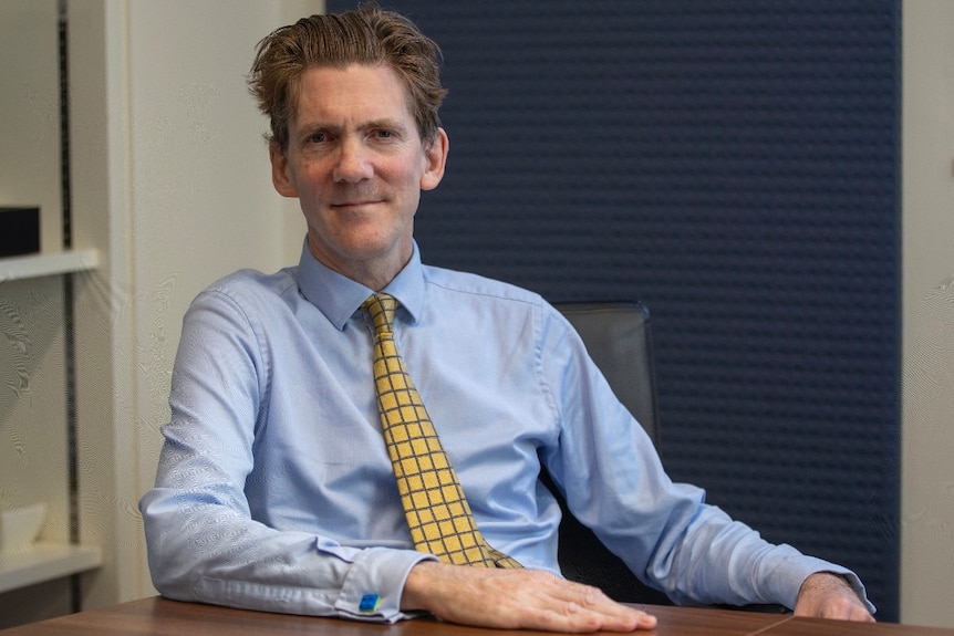 A man wearing a light blue collared shirt and yellow tie sits at a wooden desk in front of a navy curtain against a white wall.