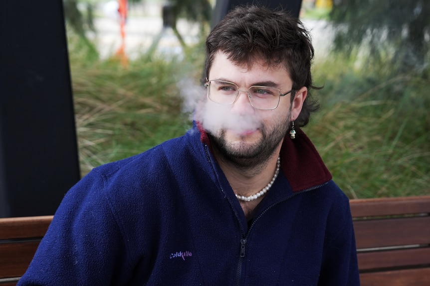 A man with short dark hair sits on an outdoor bench vaping.
