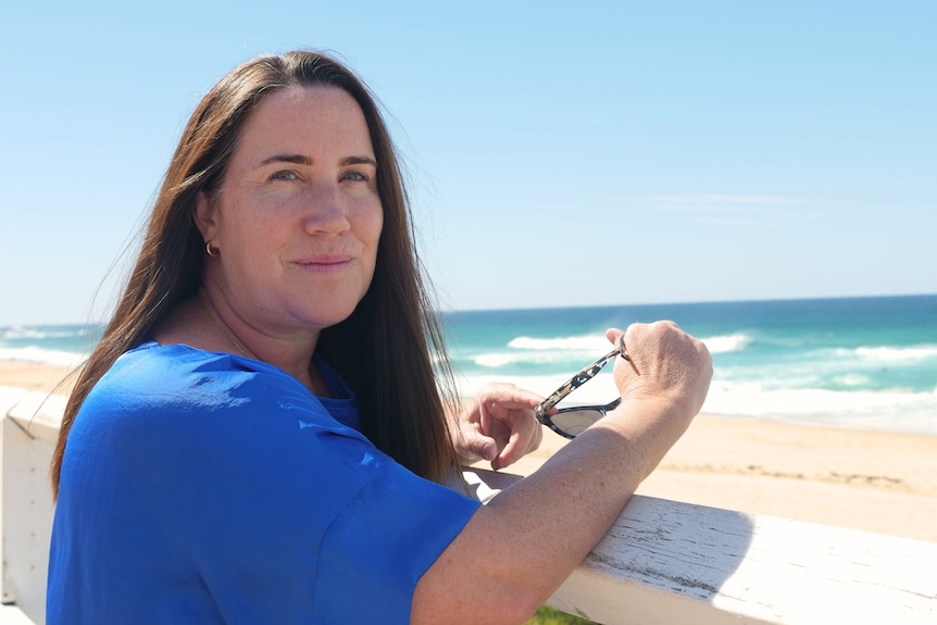 A woman looking at the camera as she leans over a fence with the ocean behind her