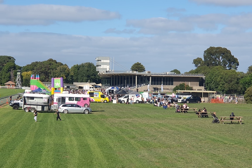 A large crowd of people, vans and a jumping castle on green grass in between a horse racing straight and a grandstand.