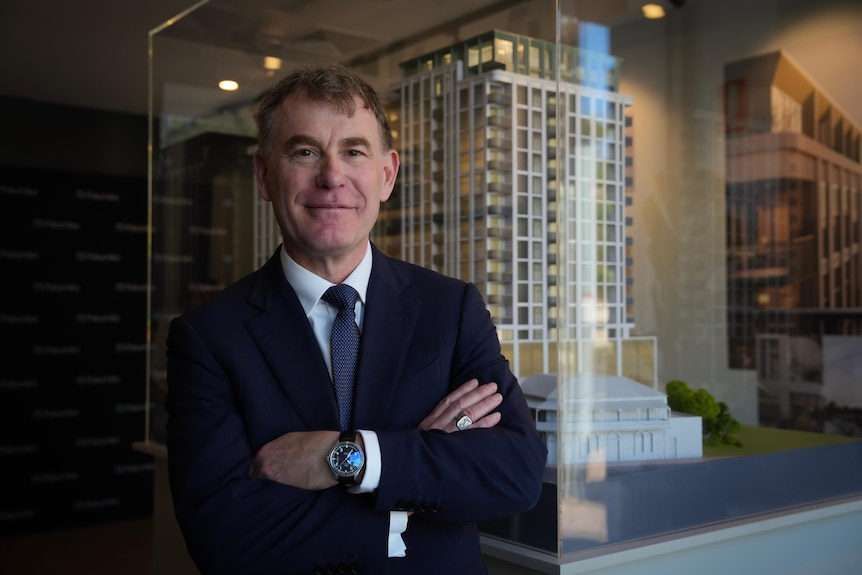 A middle-aged man in a dark suit folds his arms as he stands near a model high-rise building in a display case.