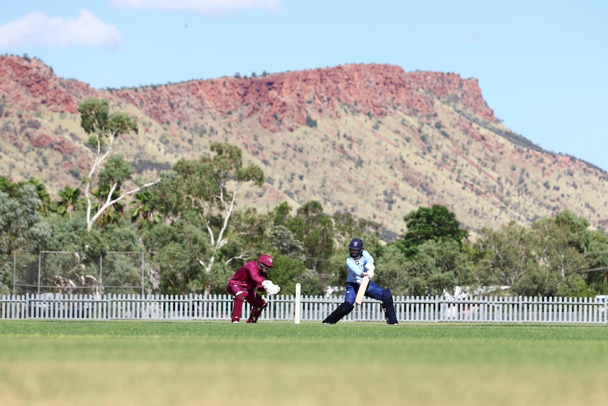 Two cricket players in action on an oval, on a sunny day, with red rocky mountains in the background.