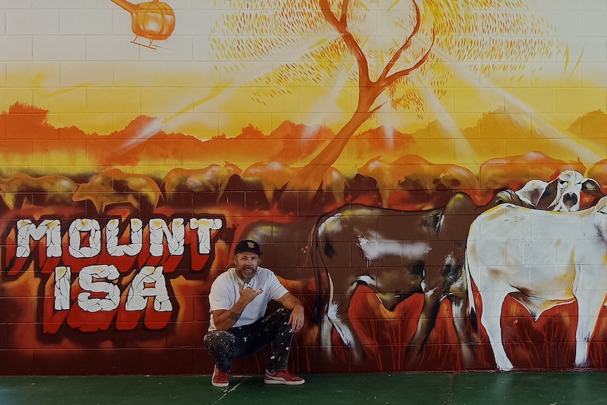 A man in a white shirt crouches next to a mural of cows with the words Mount Isa on it.