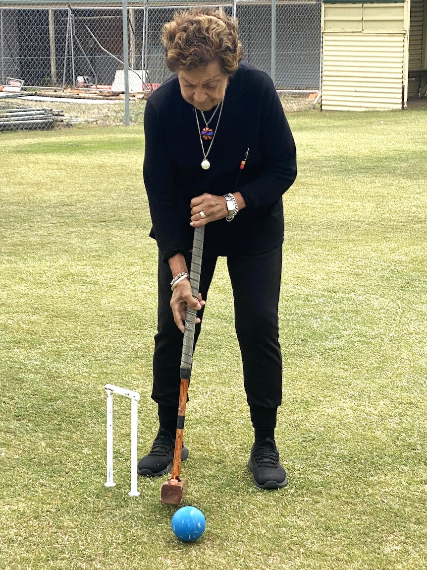 Elderly woman in black clothing with dark hair having a croquet shot with and wooden mallet, blue ball and hoop