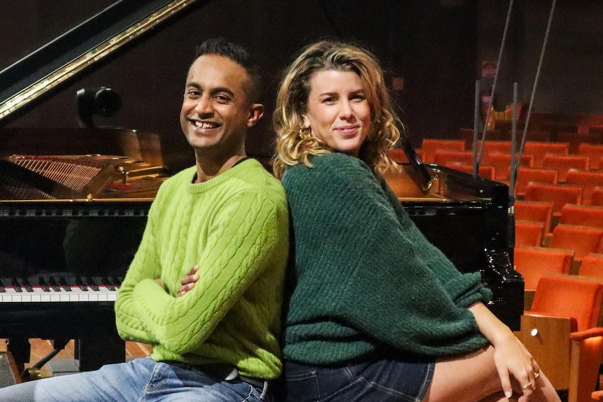 Jeremy Fernandez and Megan Burslem smile and sit back to back on a piano stool in front of a grand piano on a concert stage.