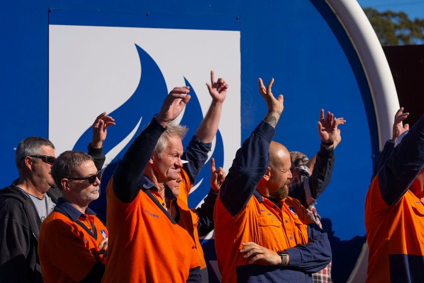 men and women in orange high vis outside a blue logo and a rusted sculpture
