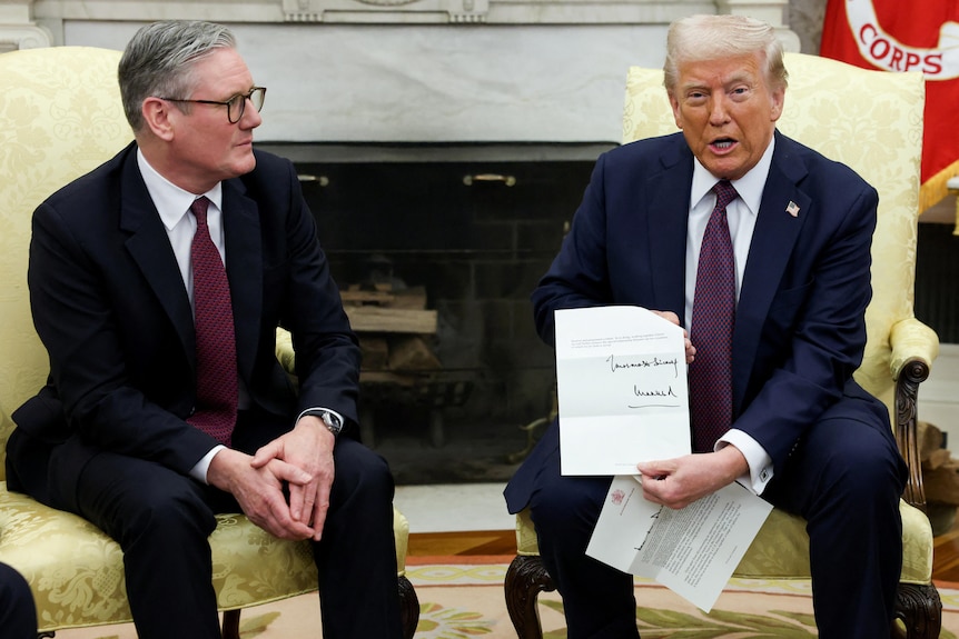 Two men in suits sit next to each other as one man holds a letter and speaks to the camera