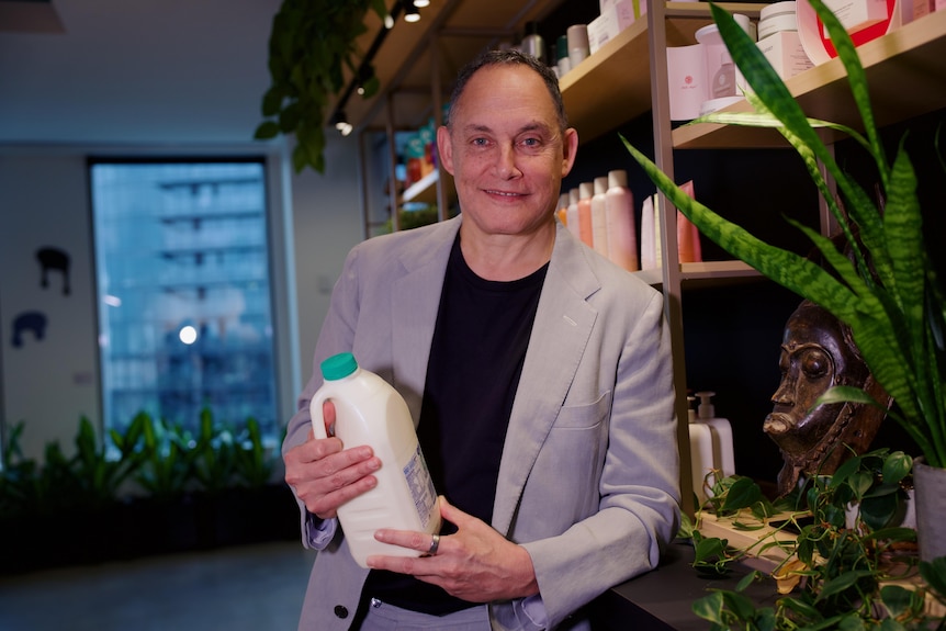 Raphael smiles, holds milk bottle, wears a light jacket, black tee, stands near shelf with plastic bottles, plant, wooden mask.