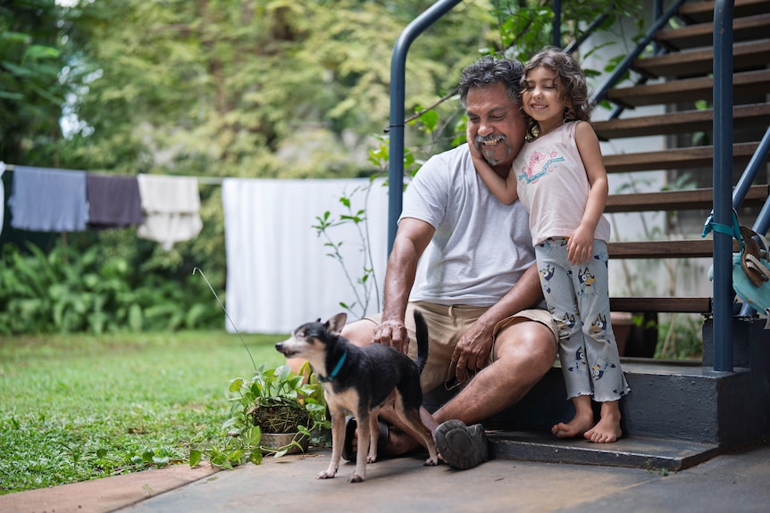 Tan man sitting on stairs outside home in backyard, young girl standing, arm around his face, smiling together at chihuahua 