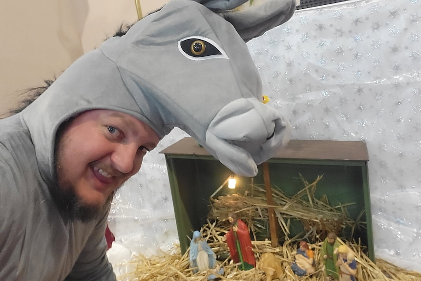 A man in a donkey costume stands in front of a nativity scene that sits on a bed of hay.