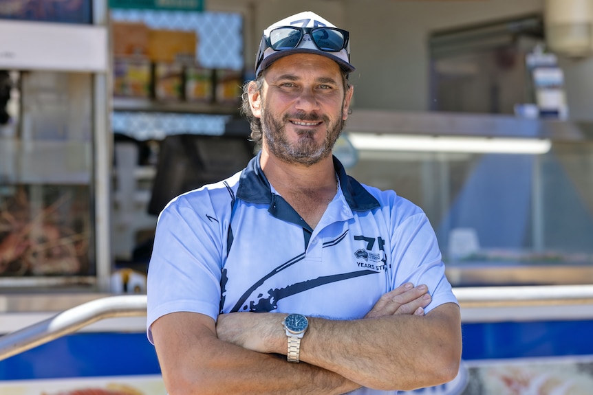A man wearing a hat stands outside a fish shop. His shirt is blue and he wears a hat. 