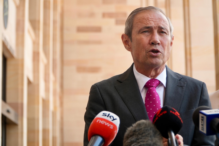 A middle-aged man with short hair wears a suit as he speaks to the media in front of a large building.