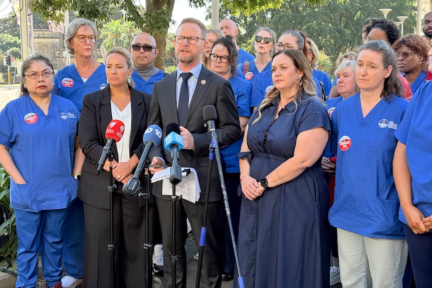 Michael Whaites at a press conference in a suit, flagged by nurses and midwives in blue uniforms.