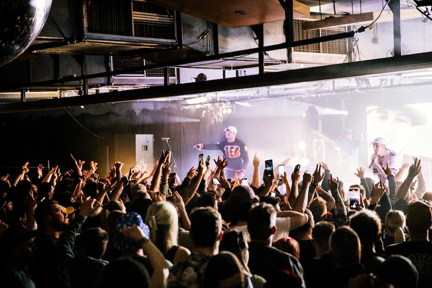 A crowd faces away from the camera and towards a stage where Bliss n Eso are performing