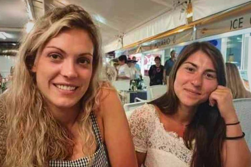 Blonde woman with dark blonde wavey hair out, seated at a table. A brunette woman sits beside her, hand  rests on her cheek