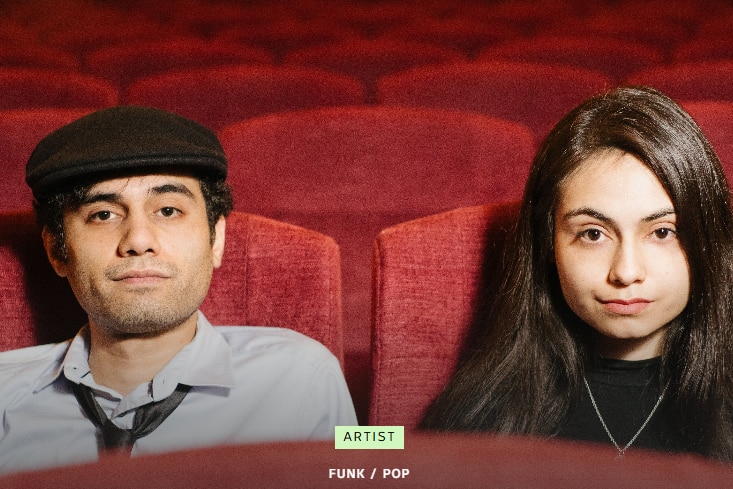 Two people sit in red cinema chairs and stare directly into the camera. Man has black paddy cap, shirt and tie. Woman in black. 