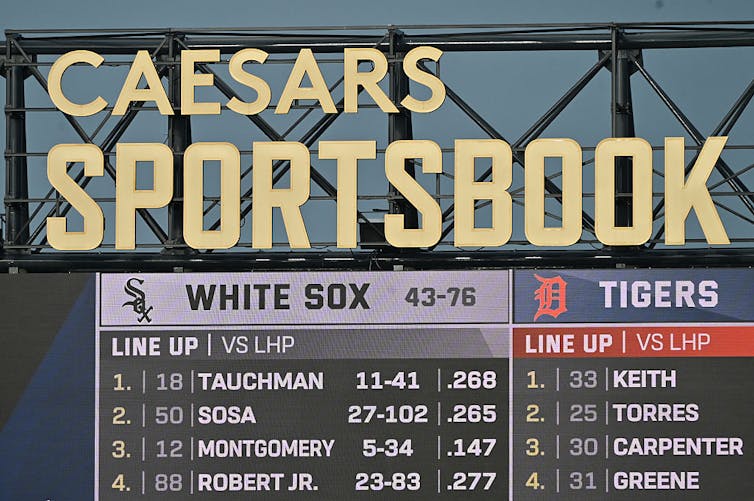 Two baseball lineups on a digital scoreboard, with a massive gold sign reading 'Caesars Sportsbook' installed above it.
