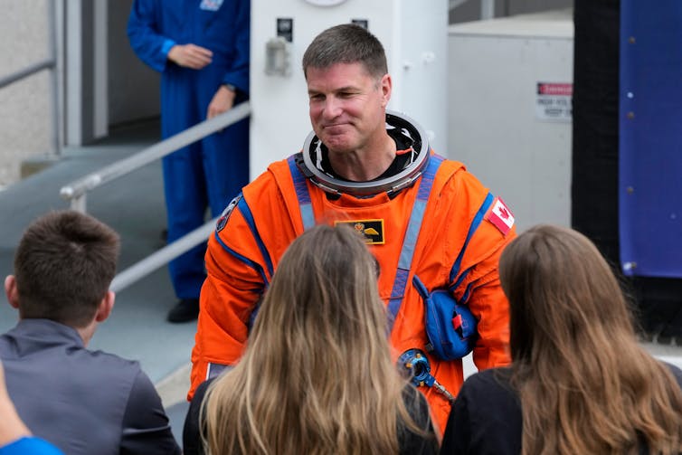 A tall man in an orange space suit smiles.