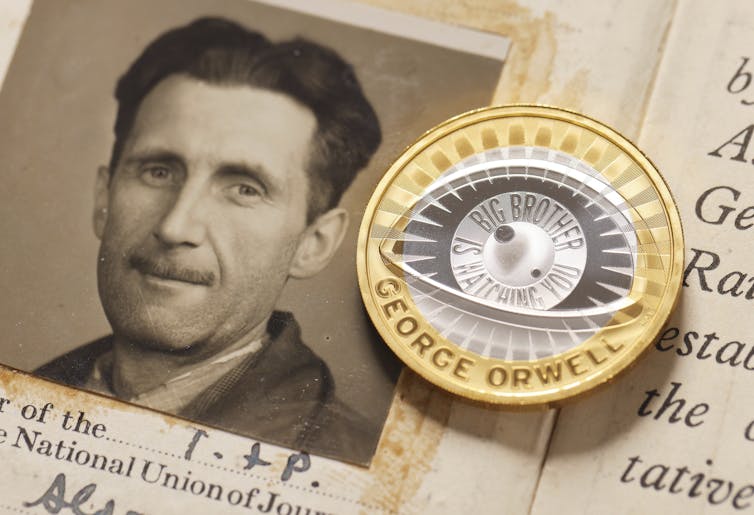 A gold-colored coin with an engraving reading 'Big Brother is Watching You' lies next to a sepia-toned portrait of a man with a thin mustache.