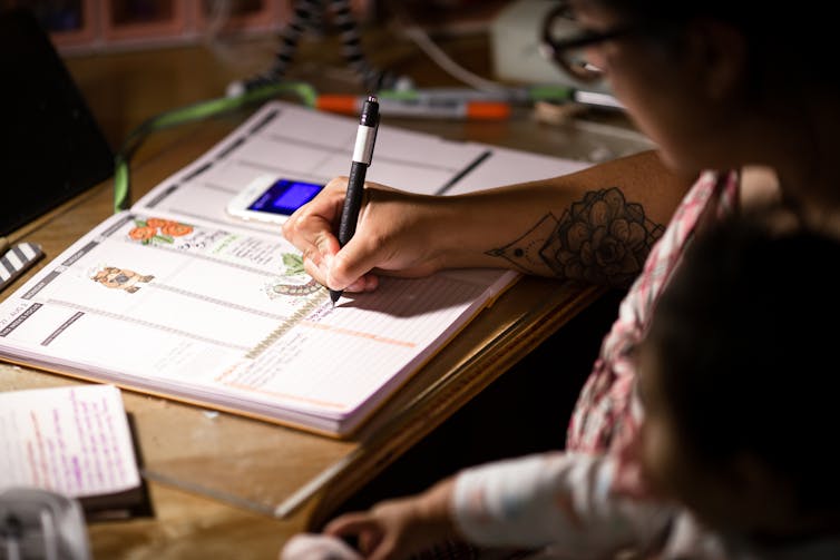 A woman holds a pen as she writes in an agenda book, sitting bent over a table.