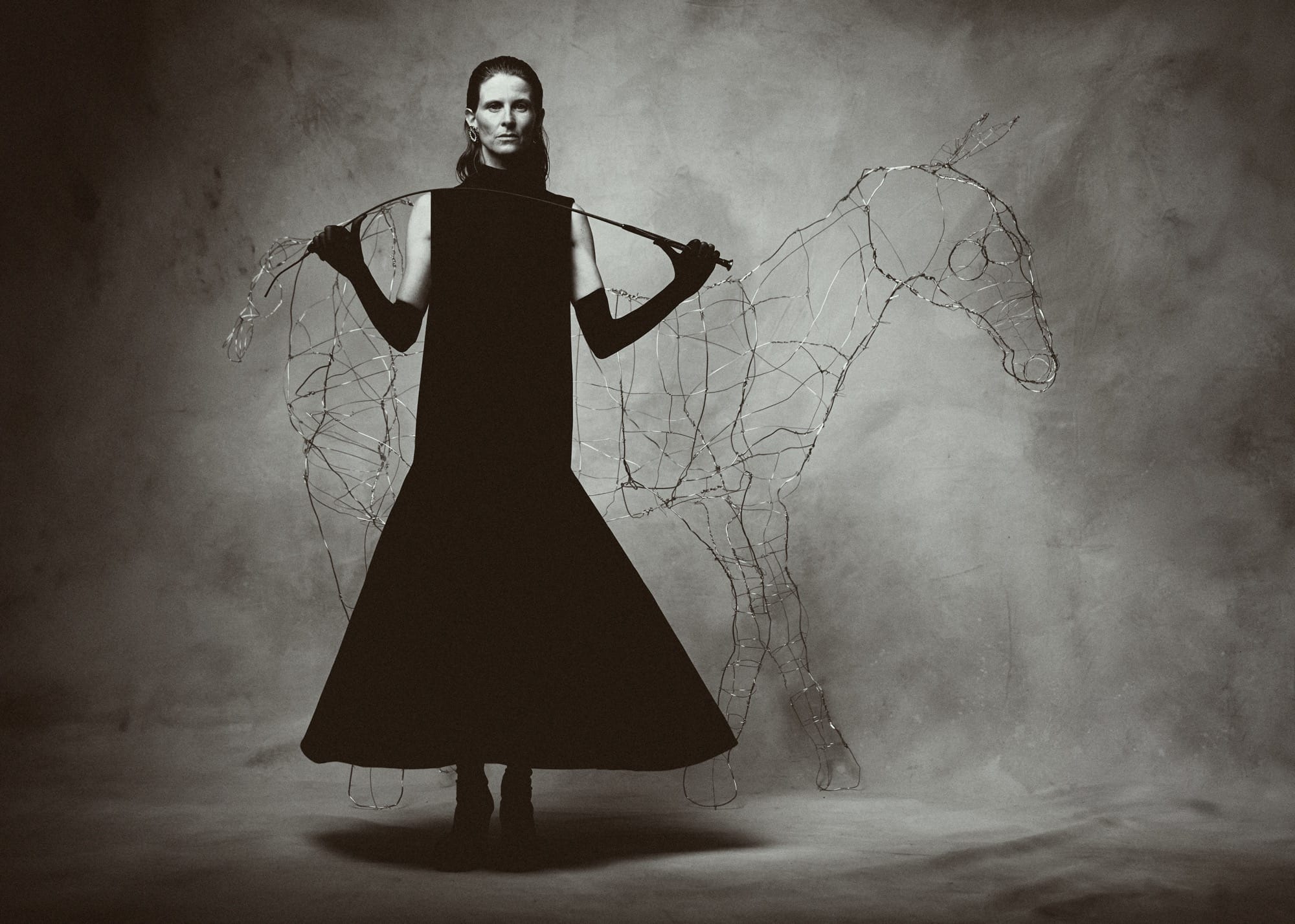 a black and white photo by Austn Fischer of a woman in a black dress with a wire horse sculpture