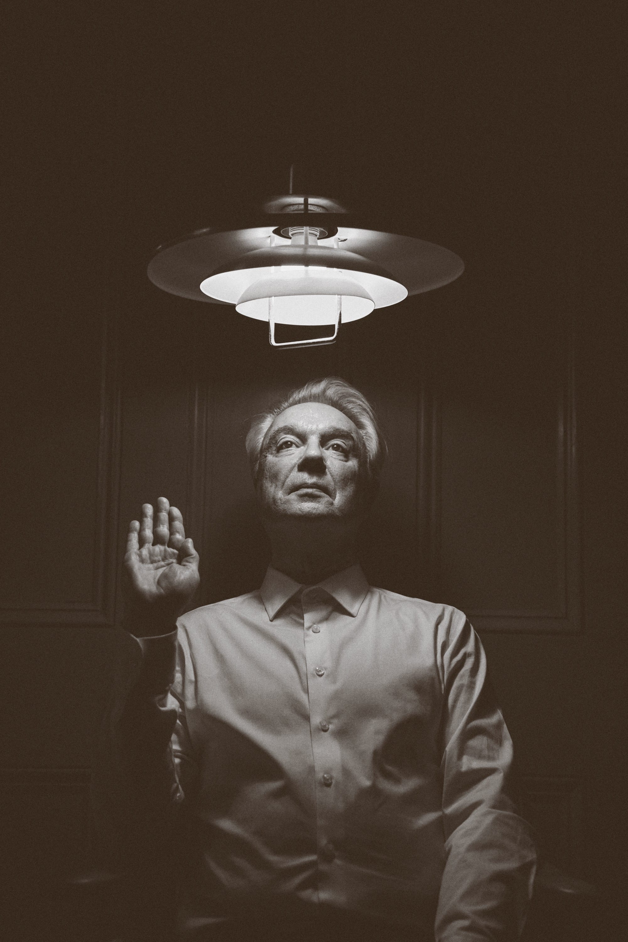 a black and white photo by Austn Fischer of david byrne underneath a light with his hand raised as if giving an oath