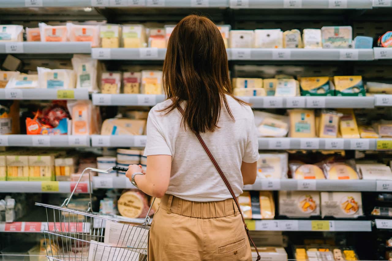 Rear view of young woman carrying shopping basket, choosing cheese, standing in front of produce aisle in supermarket