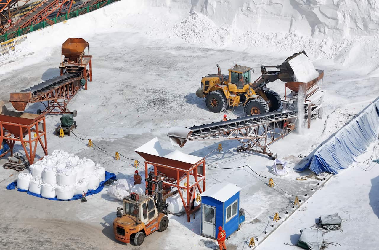 A front-end loader pours a white, crystalline substance into a processing hopper at an industrial facility, while a forklift and workers manage large bulk bags of the material nearby.