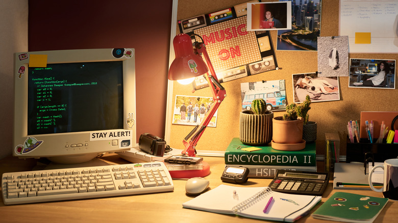 An old-school desk setup featuring a '90s-style computer setup with books, office supplies, old digital cameras, and wall decor in view.