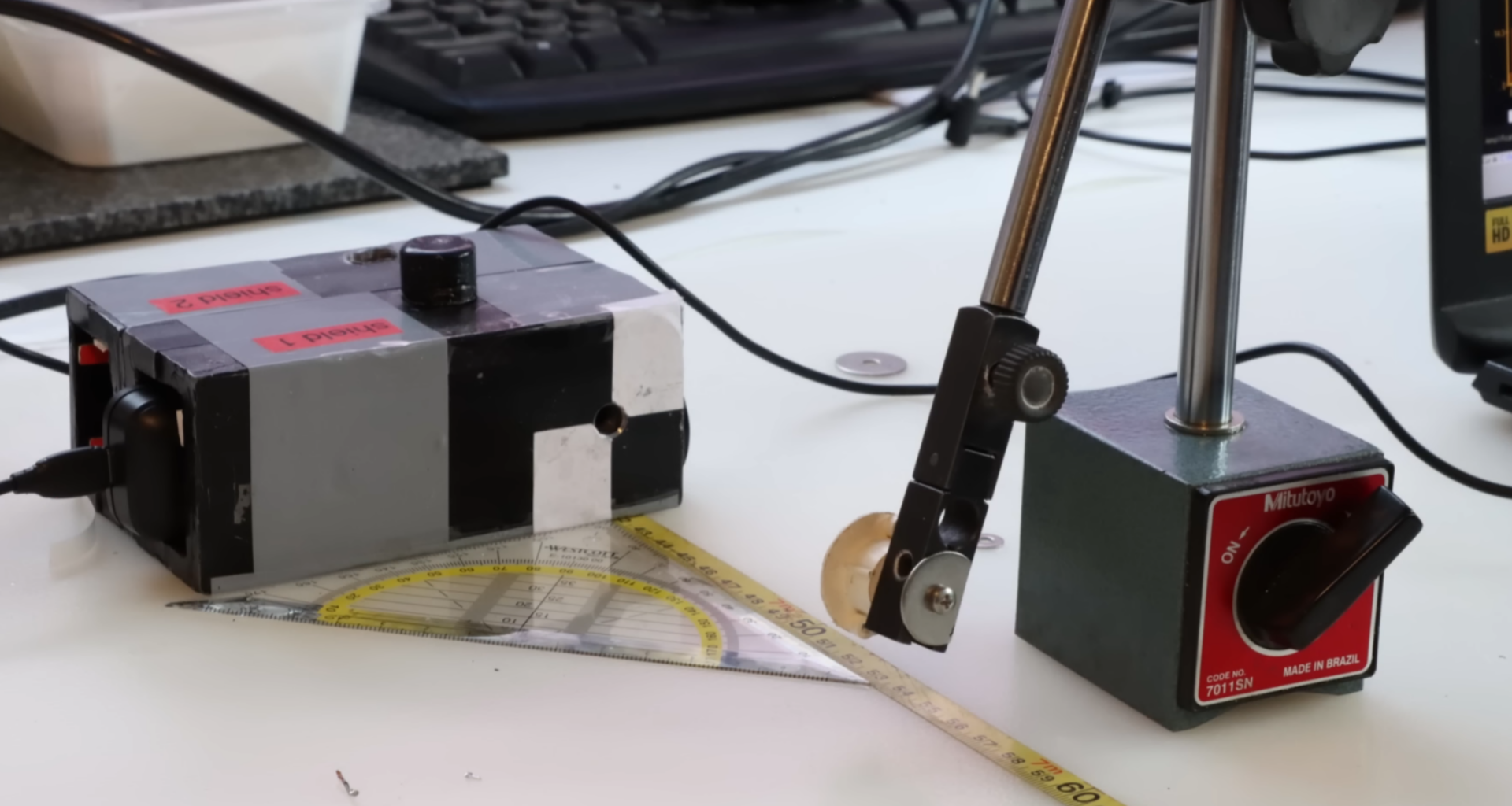 A lead box with a small aperture sits on a desk. A ruler leads away from it. A small disk on a stand is held in front of the aperture.