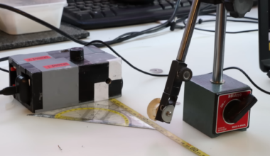 A lead box with a small aperture sits on a desk. A ruler leads away from it. A small disk on a stand is held in front of the aperture.