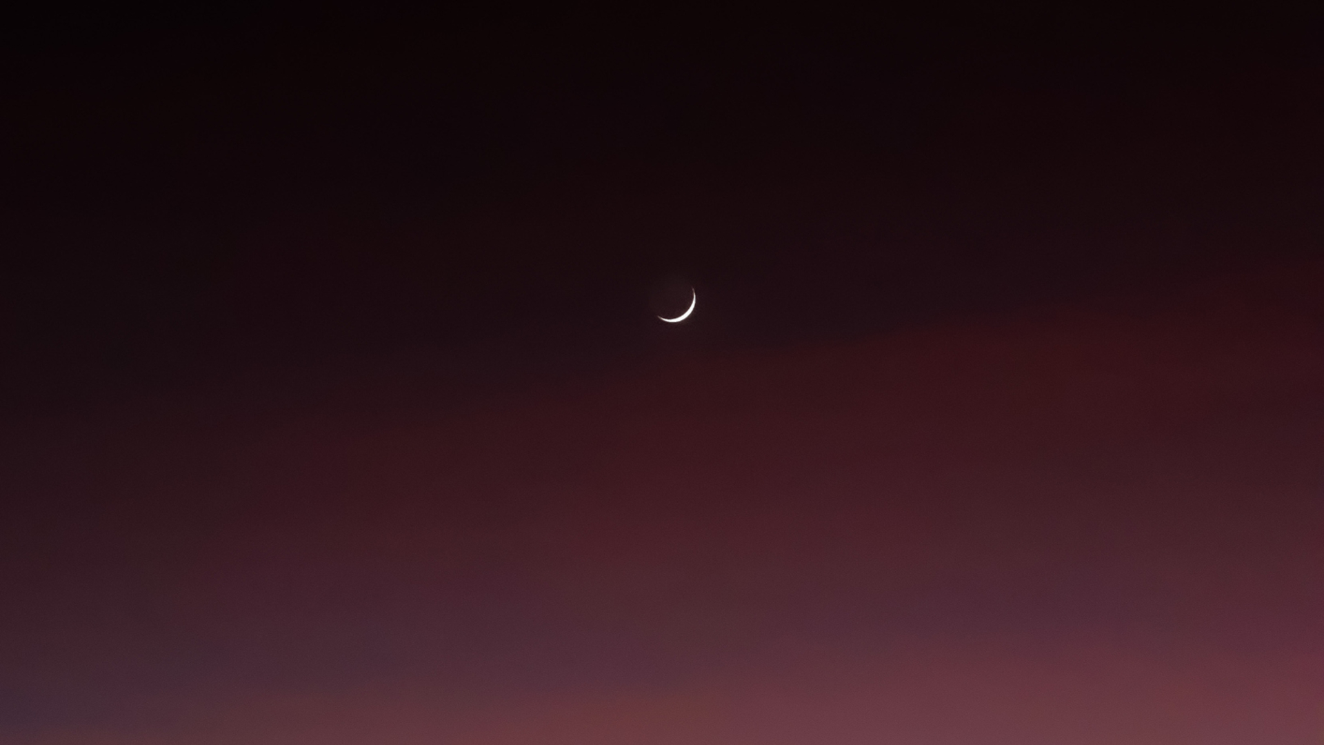 A white sliver of the moon is seen in a dark maroon night sky.