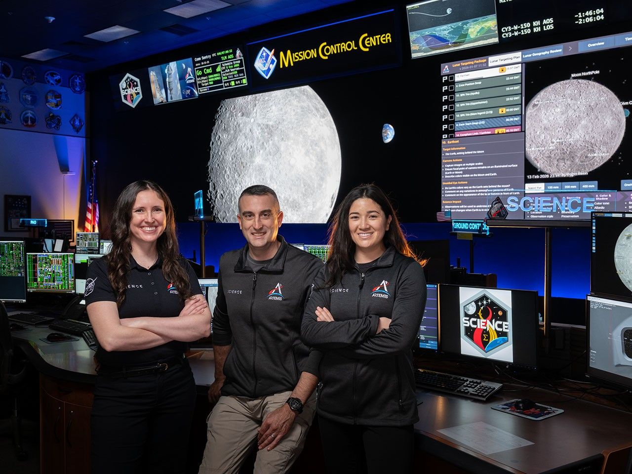 A picture of two women and a man standing at a Mission Control console desk. They are wearing shirts and jackets with the Artemis Science logo. The large display screens of Mission Control can be seen behind them, showing pictures of the Moon. The lighting in the room is dimmed and accented with blue lights.
