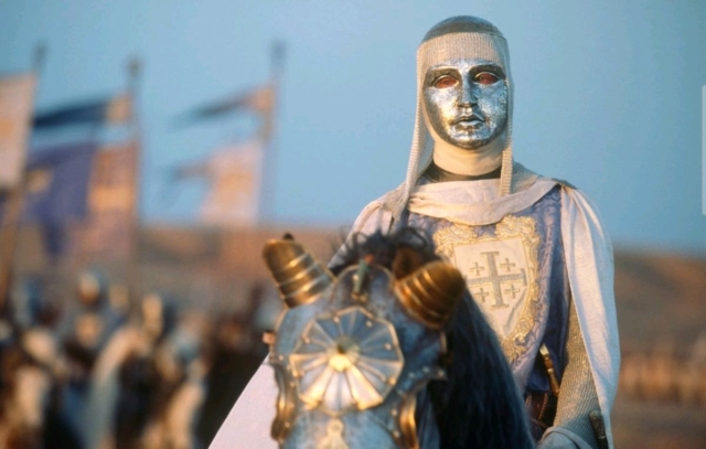 Medieval knight wearing a silver mask and armor, holding a shield, at a historical reenactment event.