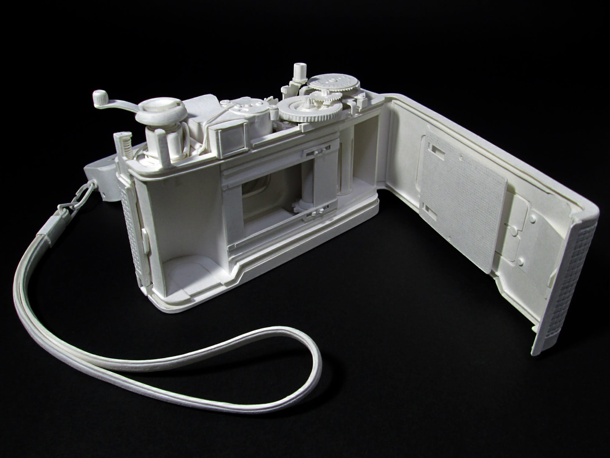 a hyperrealistic, scale sculpture of a 35mm camera made from white paper, shown open at the back