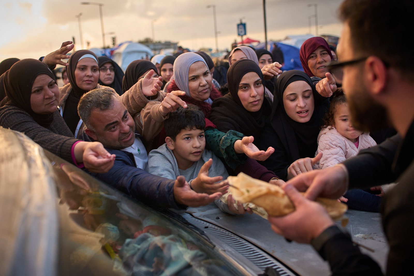 Displaced families extend their hands while waiting for donated food beside the tents they use as shelters